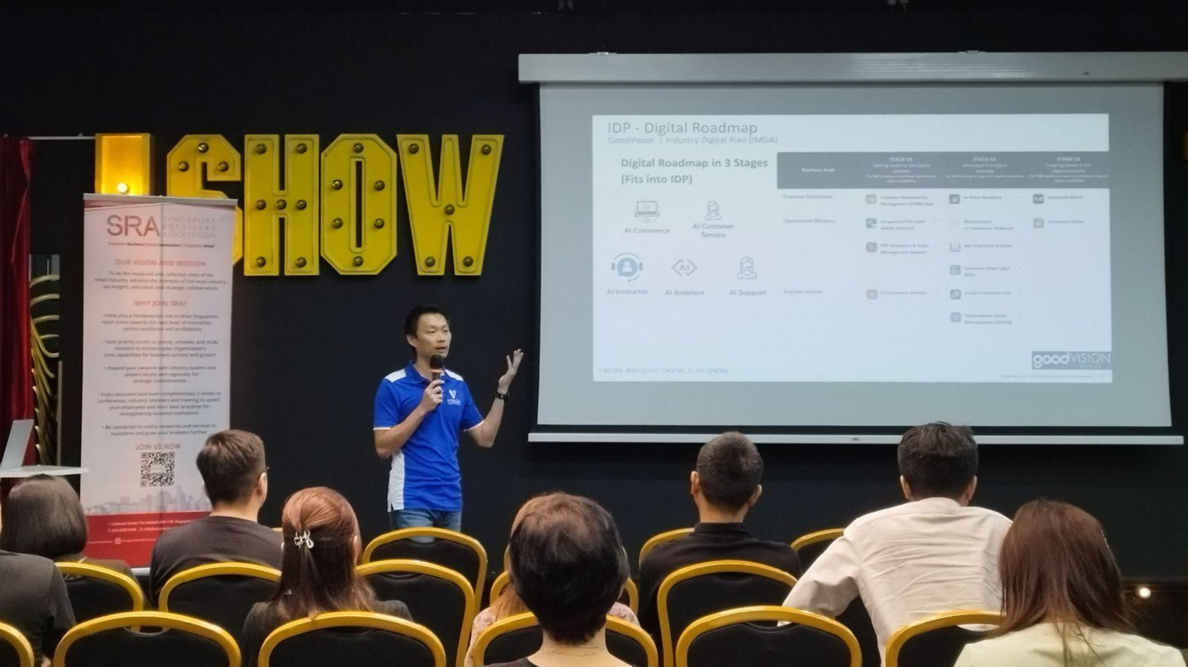 Founder and CEO of Vision Group speaks on stage about the IDP - Digital Roadmap while a slide is displayed. An assistant works on a laptop near an SRA banner. The audience listens in a room with SHOW signage and a projector screen.